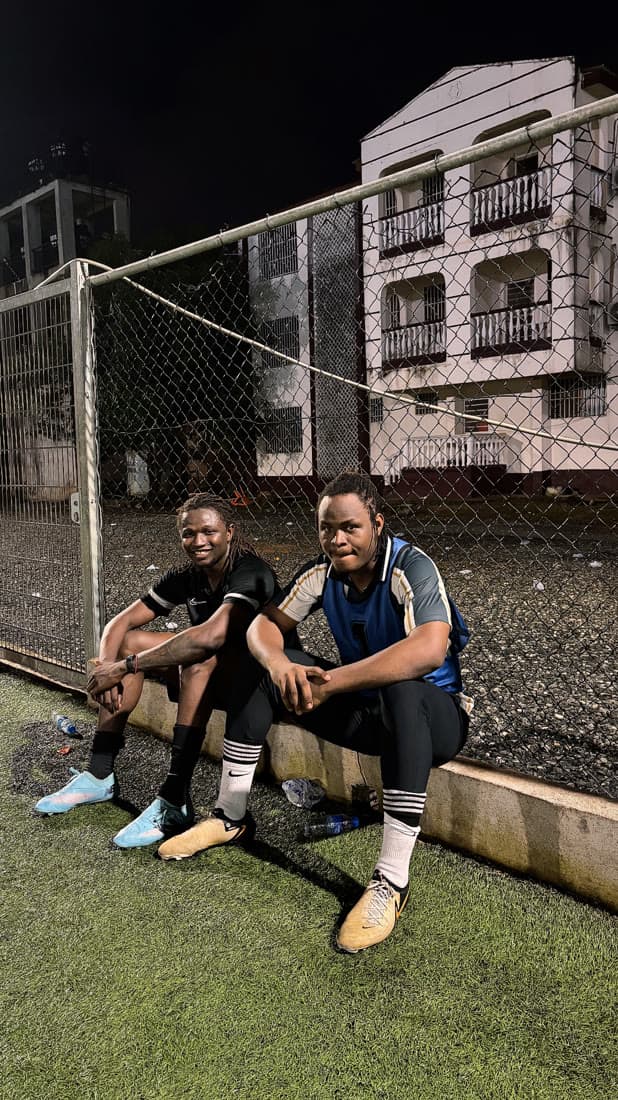 Dyte O and Minamino sitting on the pitch-side curb by the fence at night.