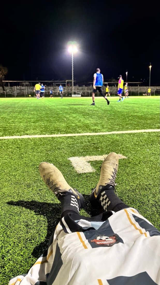 Lying on the pitch after a game, boots crossed, floodlit field beyond.